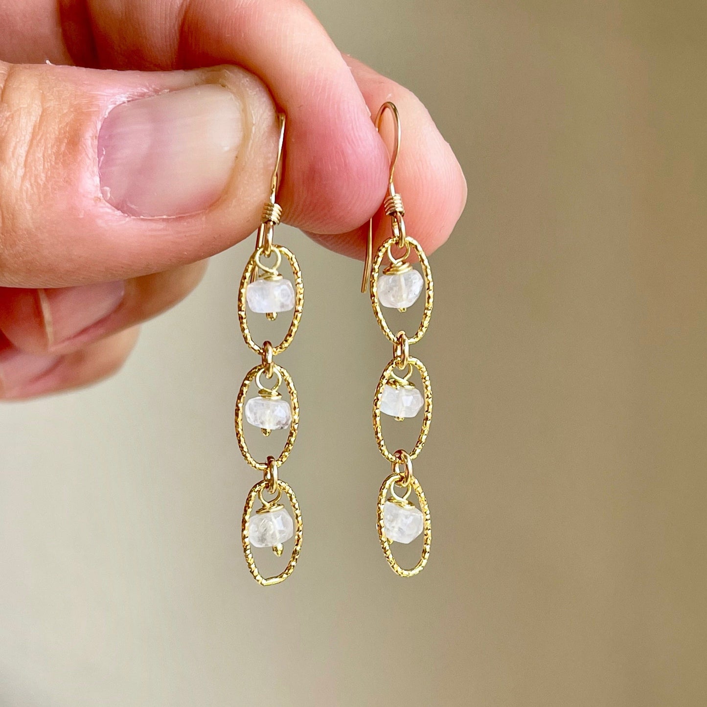 A pair of Rainbow Moonstone earrings with oval links, held in a hand against a neutral background.