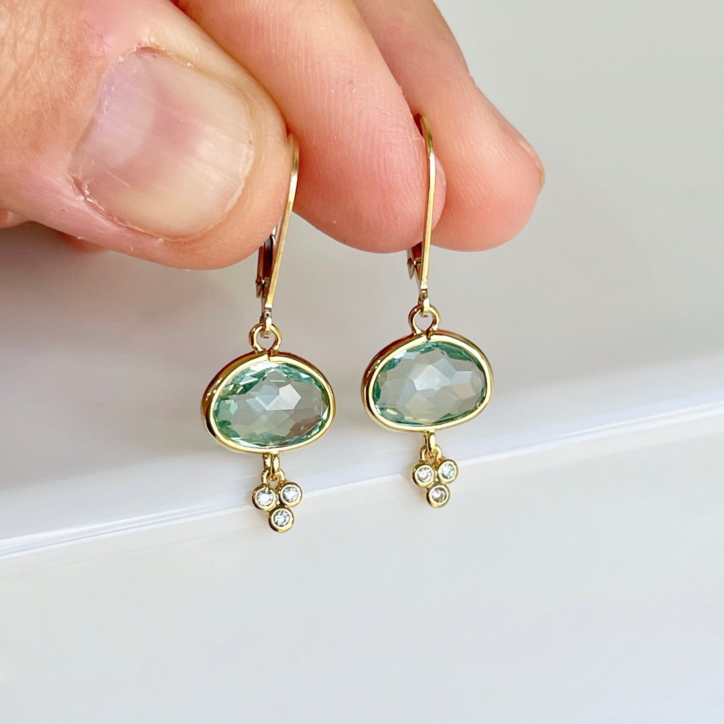 A pair of green amethyst oval earrings with gold filled or sterling silver settings, suspended from mint green prasiolite gemstones with small white topaz stones, held in a person's hand.