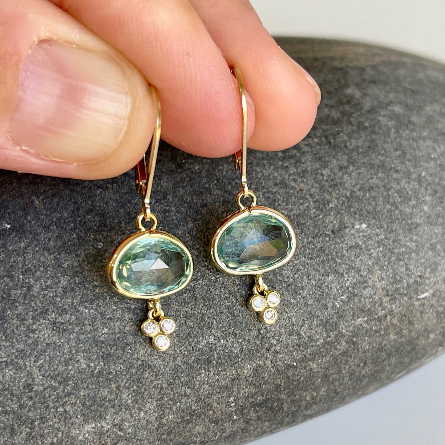 A pair of green amethyst oval earrings with gold filled or sterling silver settings, suspended from mint green prasiolite gemstones with small white topaz stones, held in a person's hand.