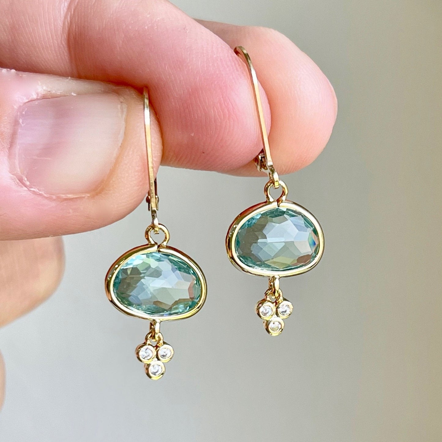 A pair of green amethyst oval earrings with gold filled or sterling silver settings, suspended from mint green prasiolite gemstones with small white topaz stones, held in a person's hand.