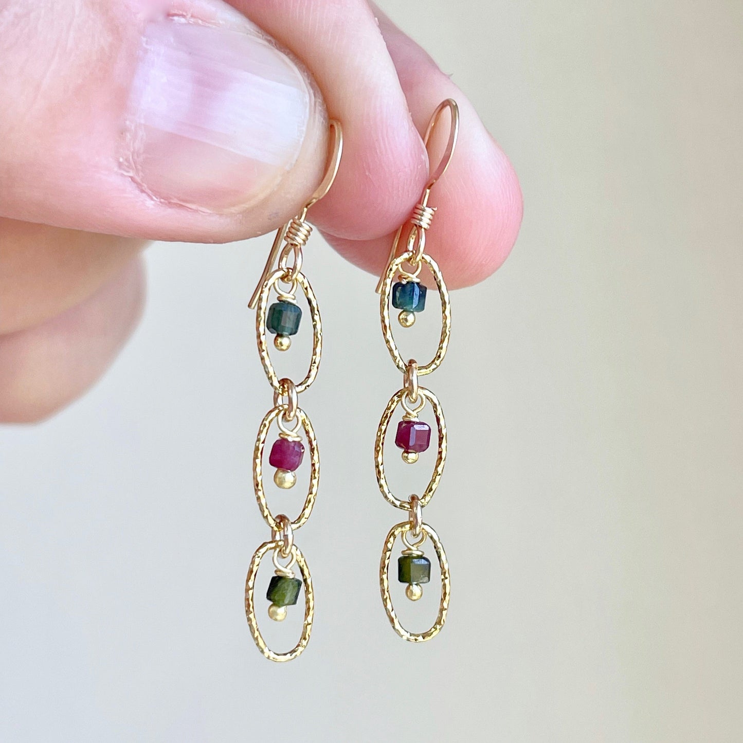A pair of watermelon tourmaline multicolor dangle earrings with gold or silver finish, held between fingers.