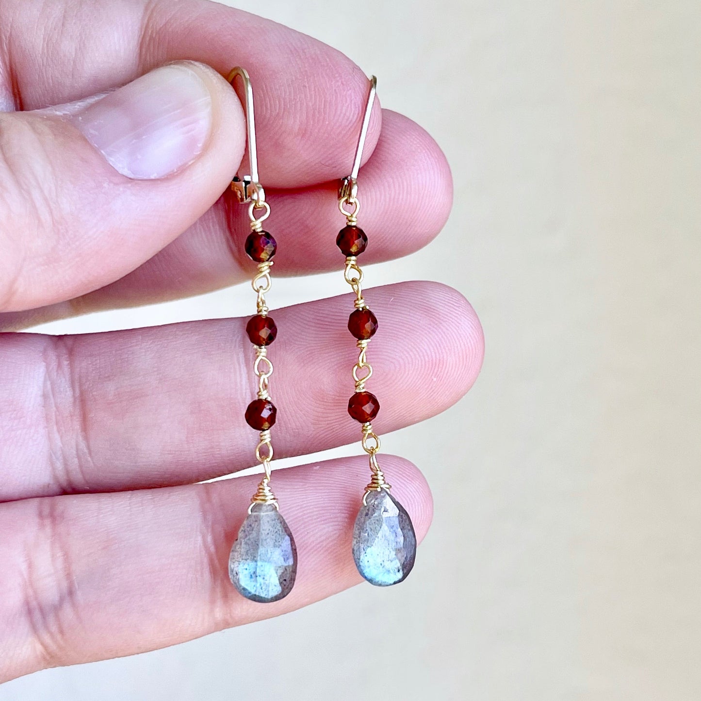 A pair of dangle earrings featuring wire wrapped Garnet stones and teardrop-shaped Labradorite stones in a person's hand.