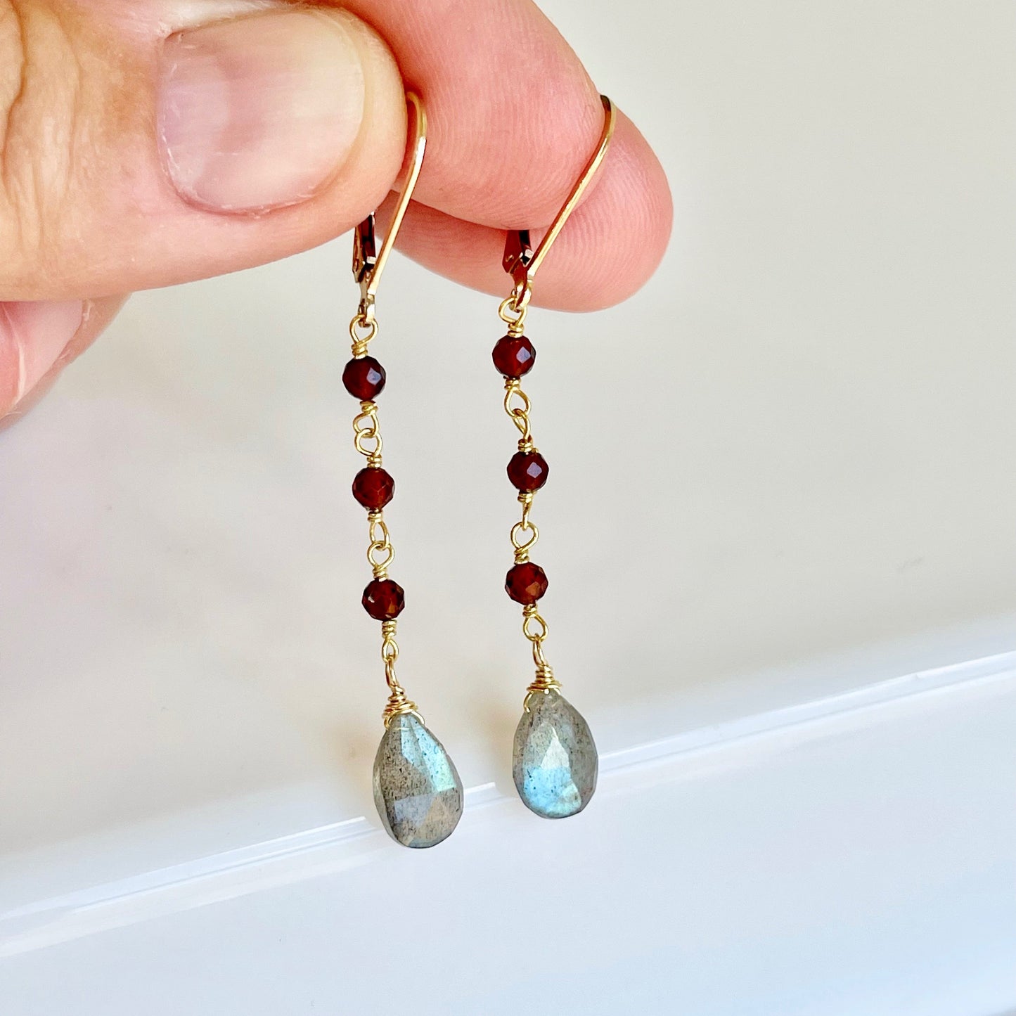 A pair of dangle earrings featuring wire wrapped Garnet stones and teardrop-shaped Labradorite stones in a person's hand.