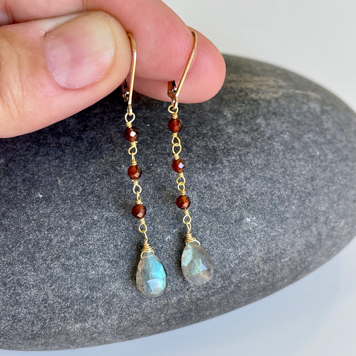 A pair of dangle earrings featuring wire wrapped Garnet stones and teardrop-shaped Labradorite stones in a person's hand.