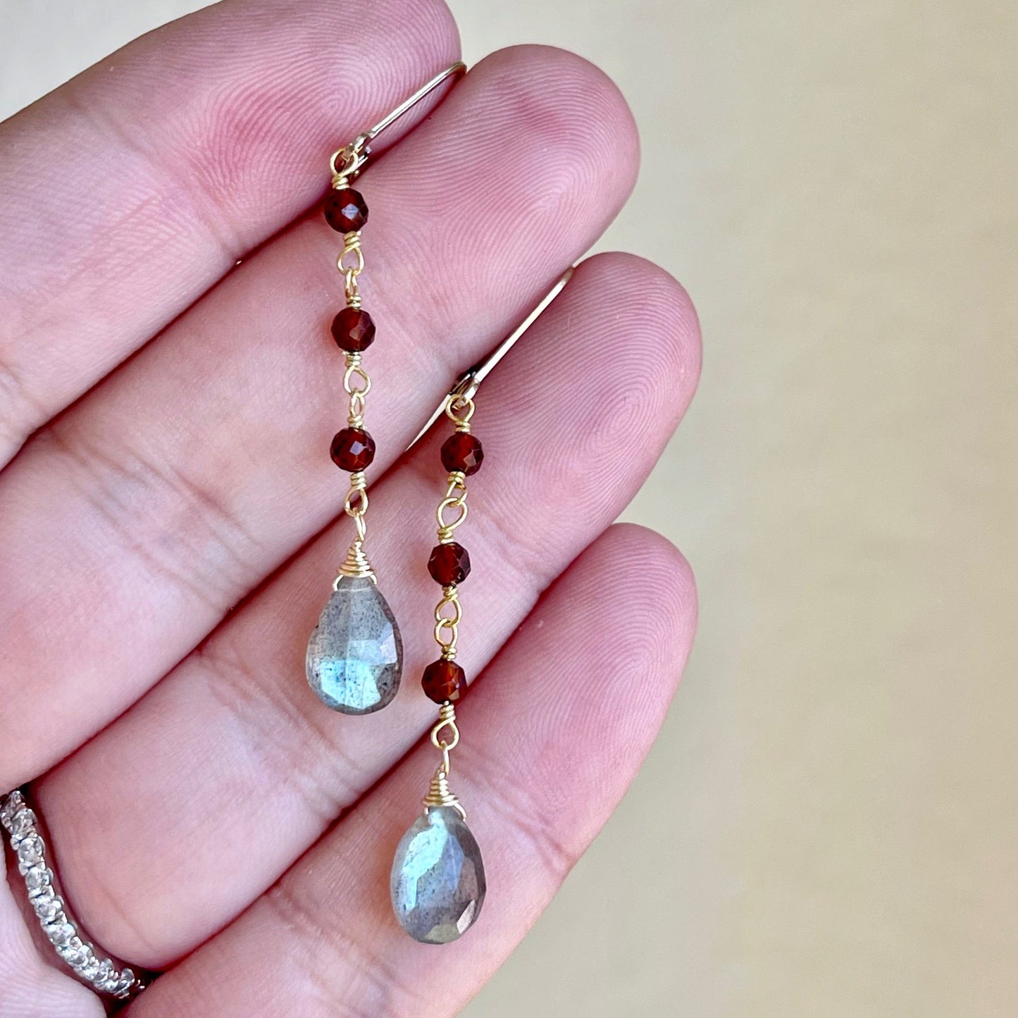 A pair of dangle earrings featuring wire wrapped Garnet stones and teardrop-shaped Labradorite stones in a person's hand.