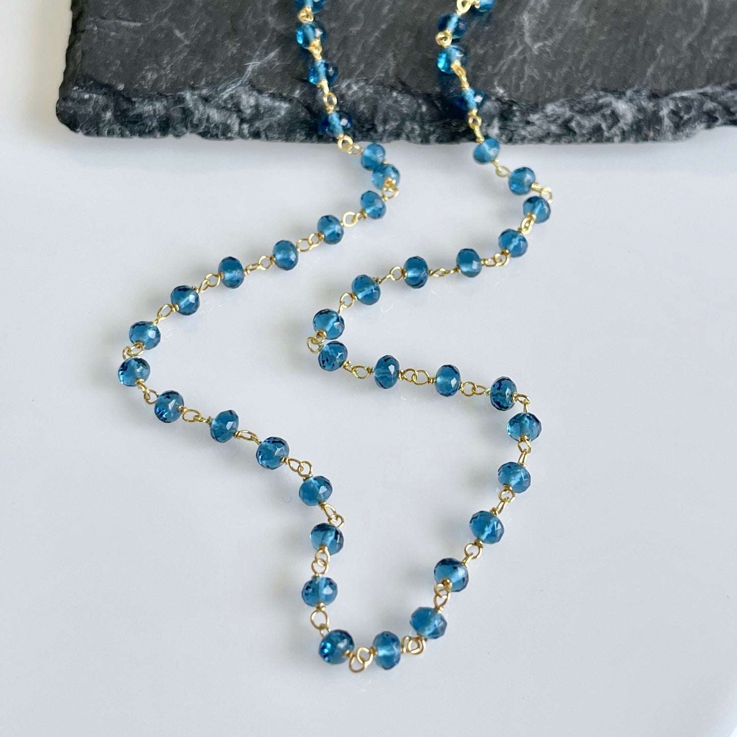 A dainty necklace with flawless 4mm micro faceted London Blue Topaz beads, wire wrapped in gold filled or sterling silver, laid out on a white surface.
