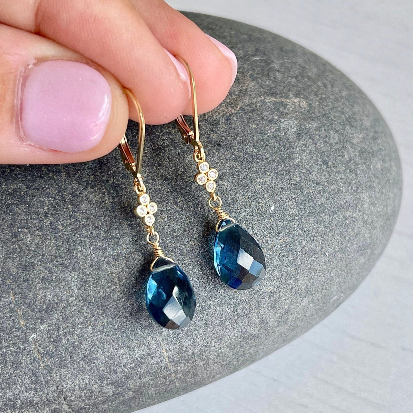 A pair of dark blue topaz teardrop dangle earrings held in a person's hand, with a gold or silver finish and a small white topaz connector between each earring and the hand.