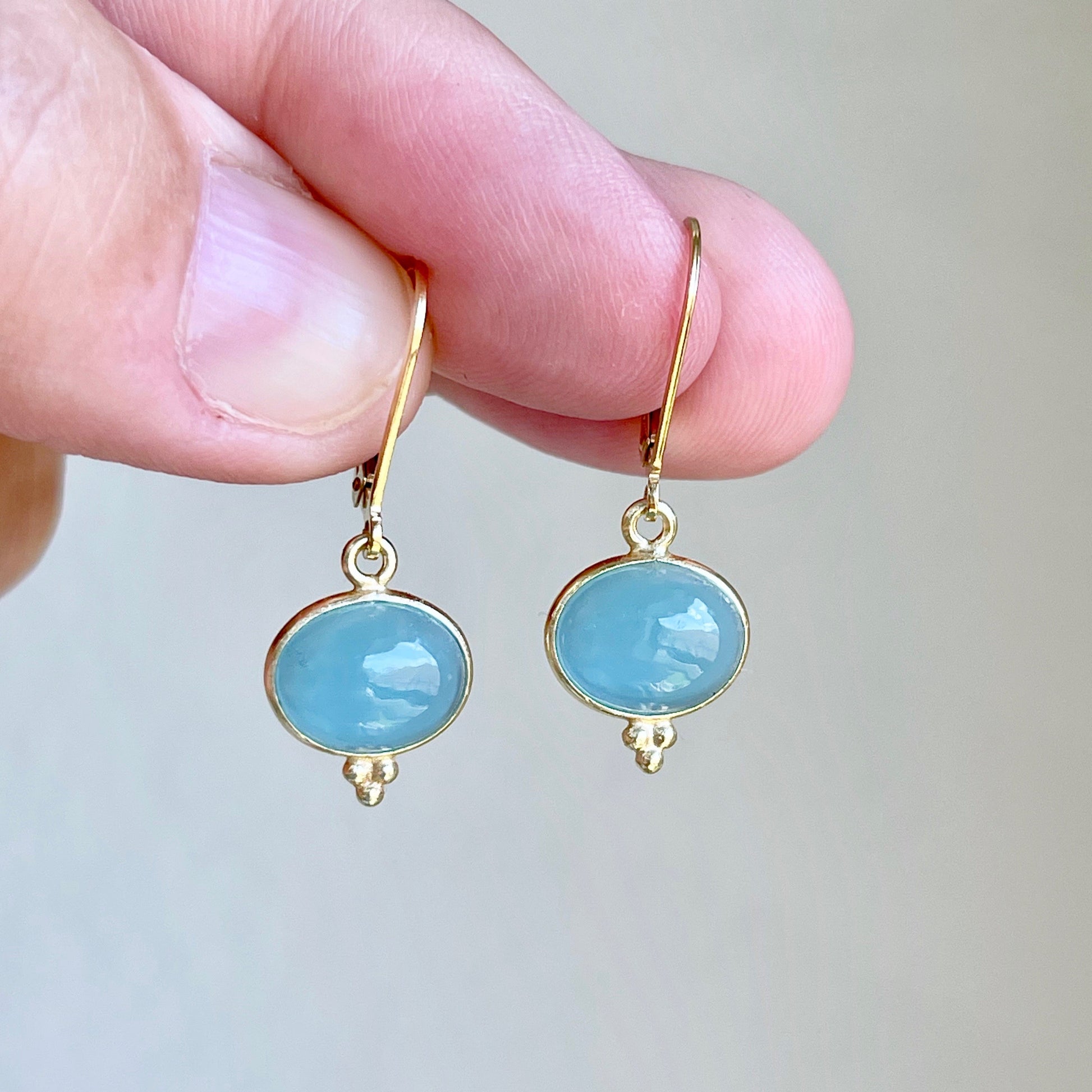 A pair of milky blue aquamarine smooth oval earrings with gold or silver settings, held in a person's hand.