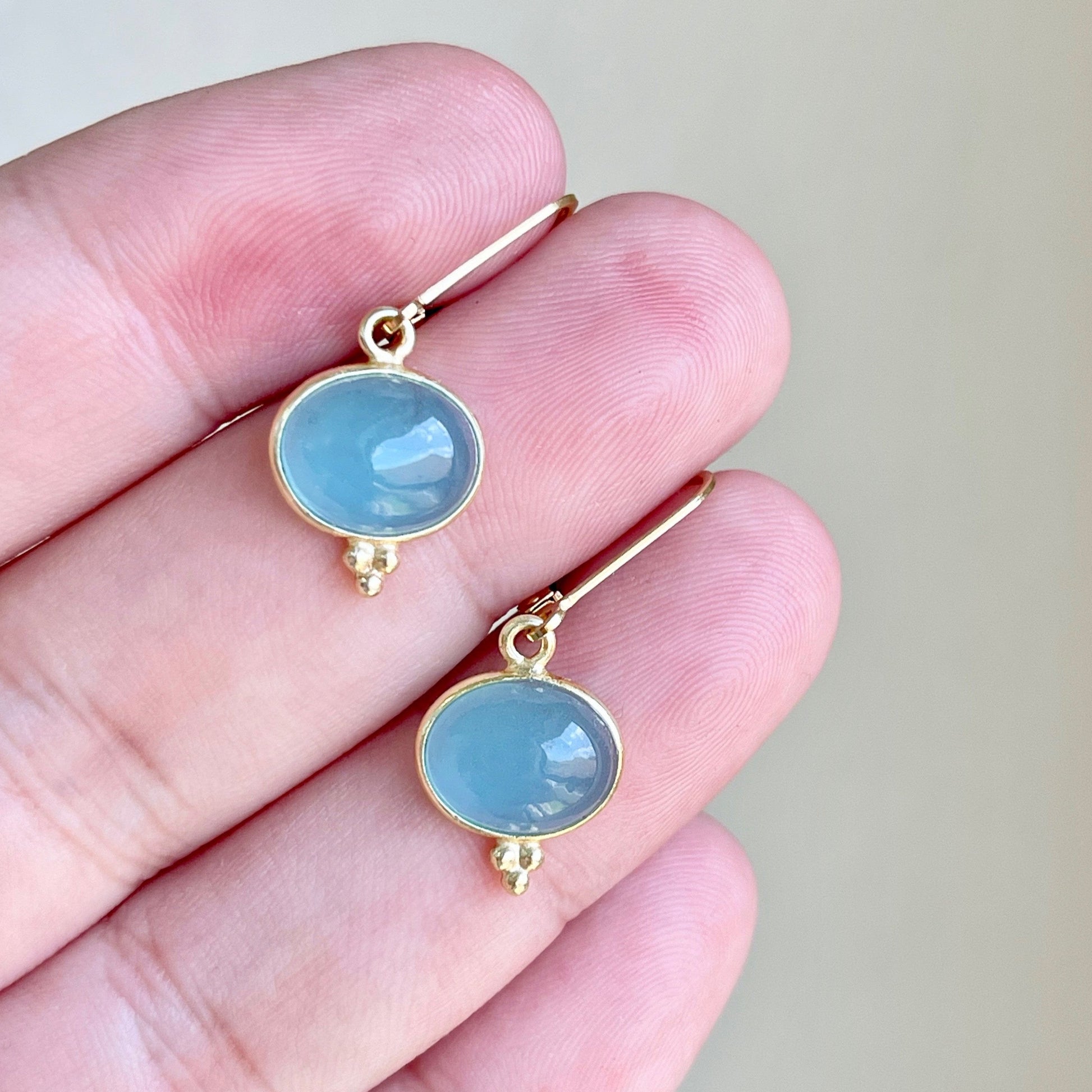 A pair of milky blue aquamarine smooth oval earrings with gold or silver settings, held in a person's hand.