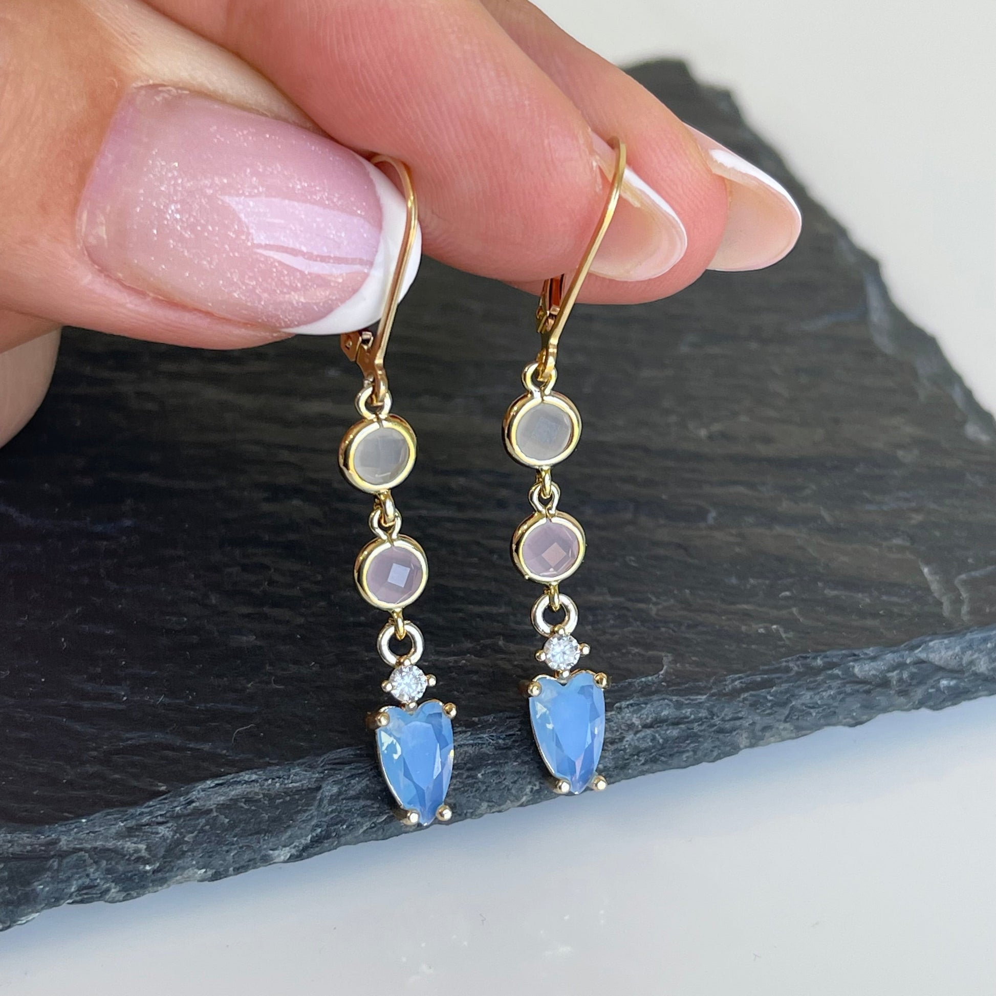 Gold earrings with blue gemstones held by a hand on a dark stone surface.